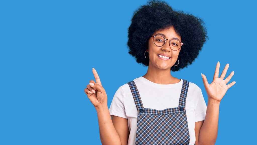 カジュアルな服を着た若いアフリカ系アメリカ人の女の子が、自信に満ちた幸せな笑顔を浮かべながら、指で6の数字を示して上を指しています。