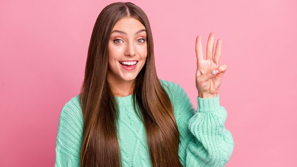 3本の指と歯を見せて微笑む魅力的な人物の肖像画。ピンク色の背景にターコイズブルーのプルオーバーが描かれている。