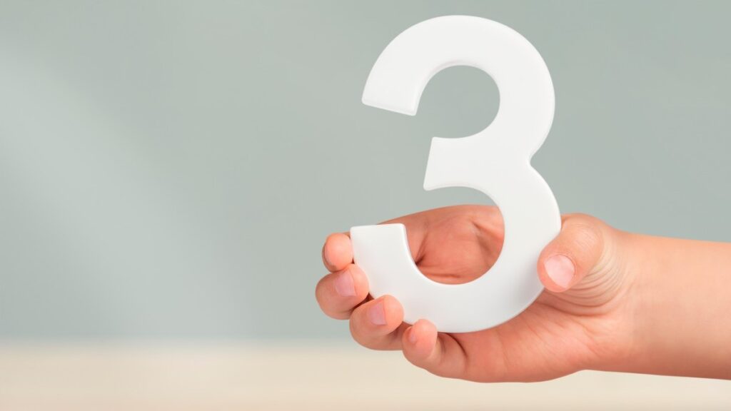 手に数字の3。ぼかしの背景にコピースペースのある白い数字の3を持つ手。数字の3のコンセプト。3%、誕生日3年、小学3年生、トリプル。
